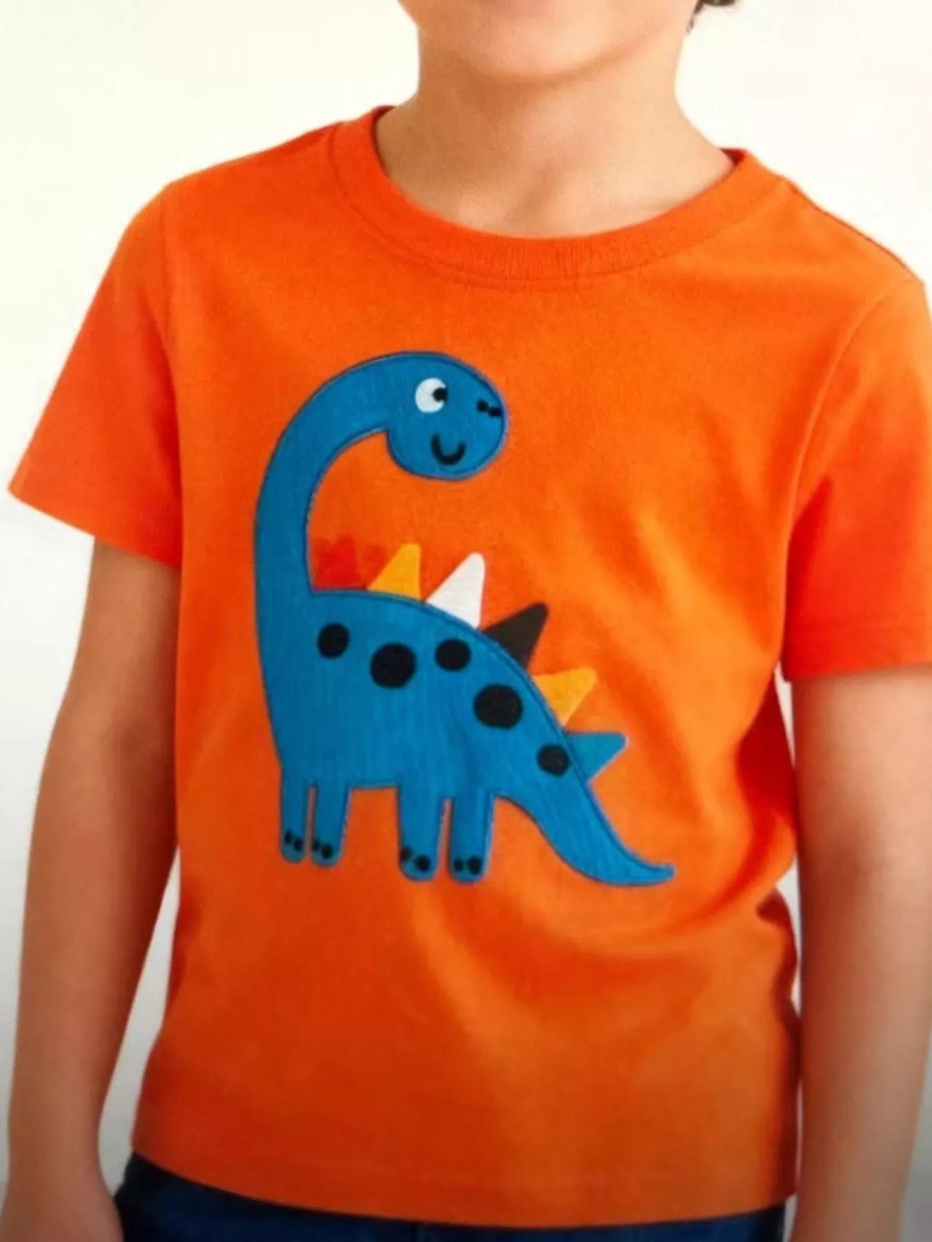 Front view of an orange children's tee with a large blue dinosaur applique and colourful back spikes