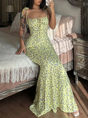 Model seated on bed taking mirror selfie in yellow floral print bodycon maxi dress with flared hem