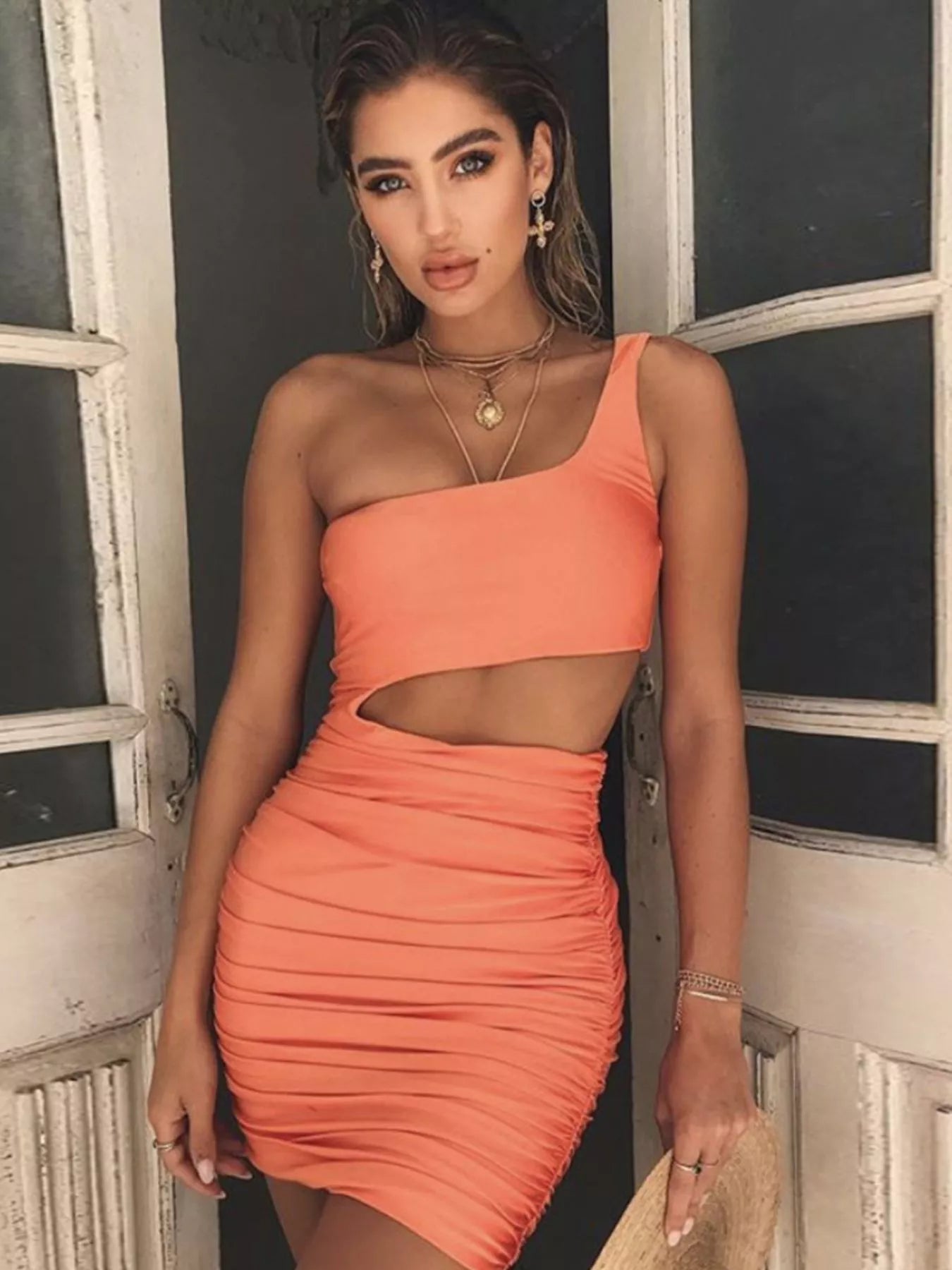 Orange powder one shoulder cutout ruched bodycon mini dress worn front view in a doorway holding a straw hat