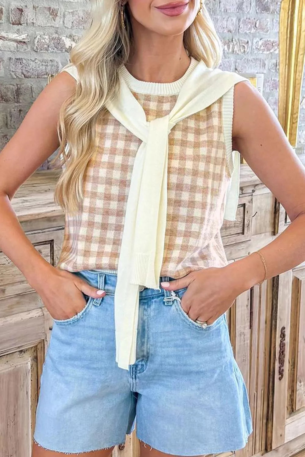 Front view of a tan gingham sleeveless sweater vest with a cream sweater draped around the neck