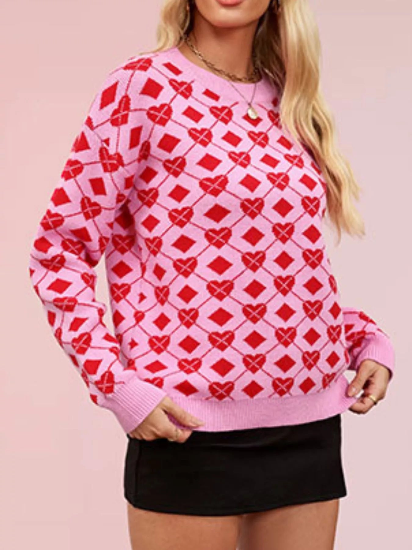 On-model front view of a pink knit sweater with red heart argyle pattern, hands at hem