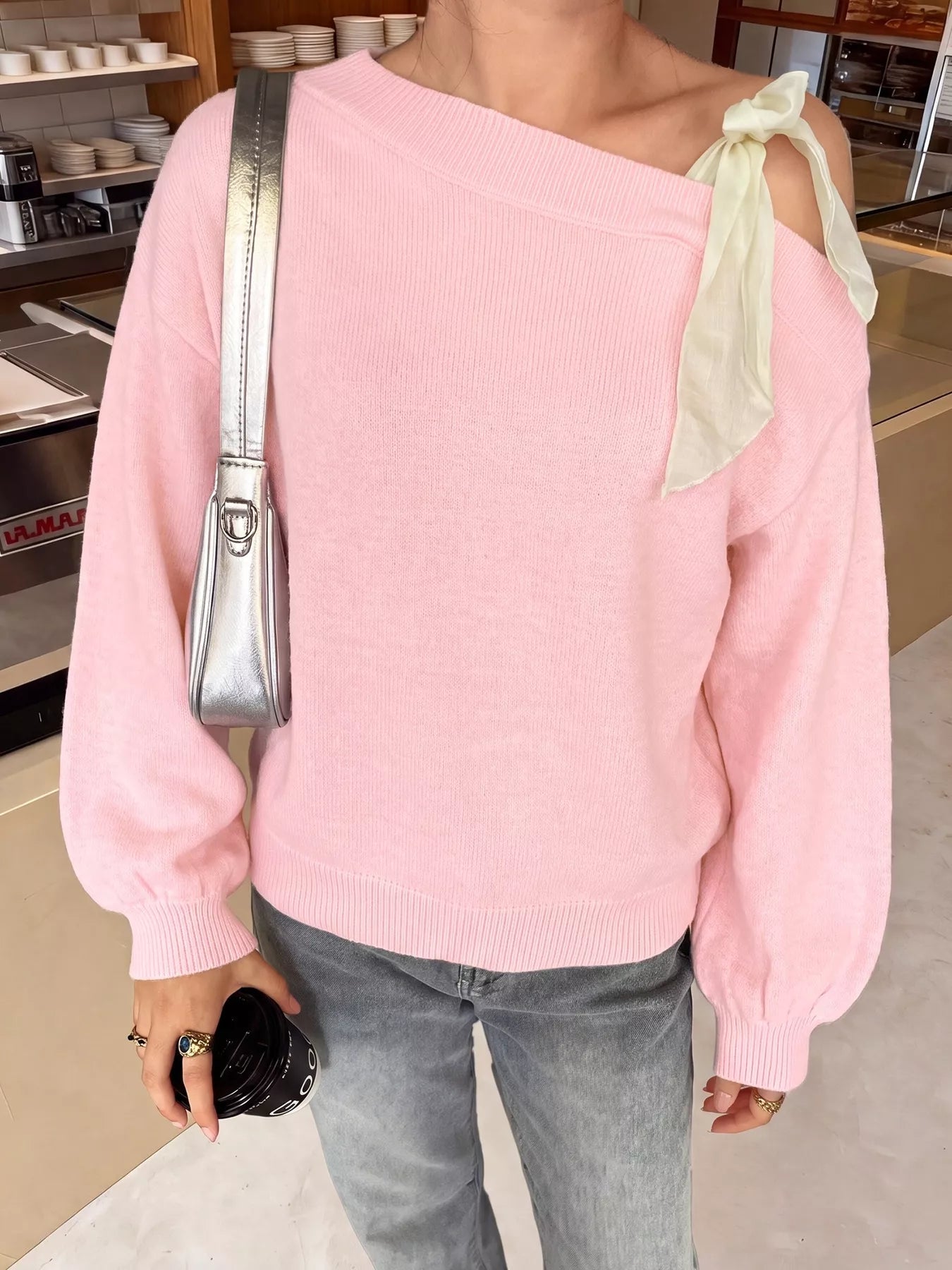 Front view of a pink one-shoulder sweater with a cream ribbon bow, shown indoors