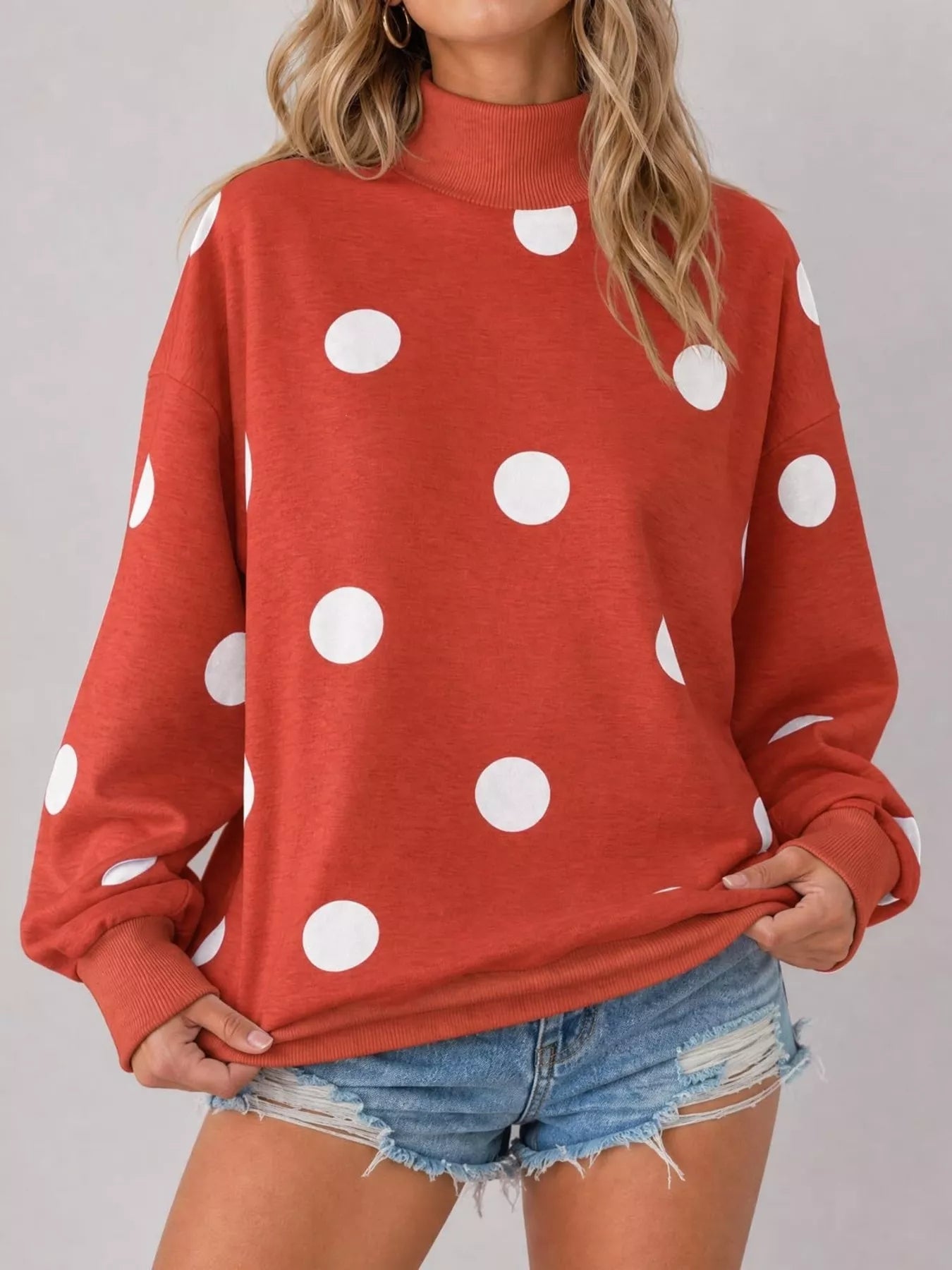 Front studio view of red mock neck oversized sweatshirt with white polka dots holding hem