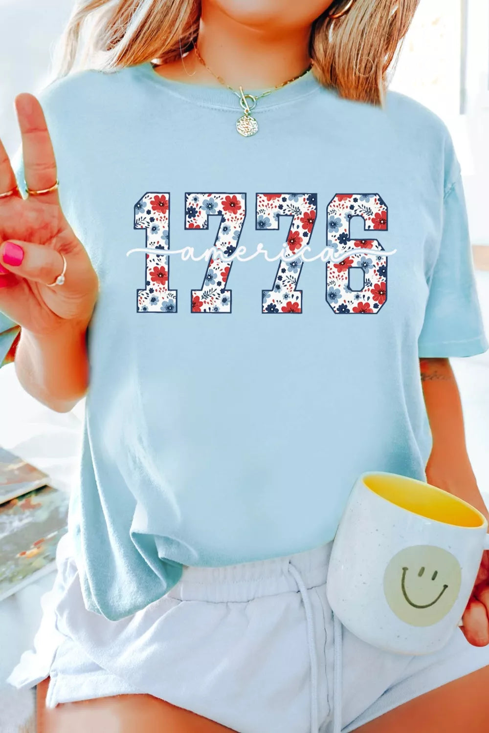 Front lifestyle view of light blue relaxed T-shirt featuring floral “1776” and cursive “America” lettering.