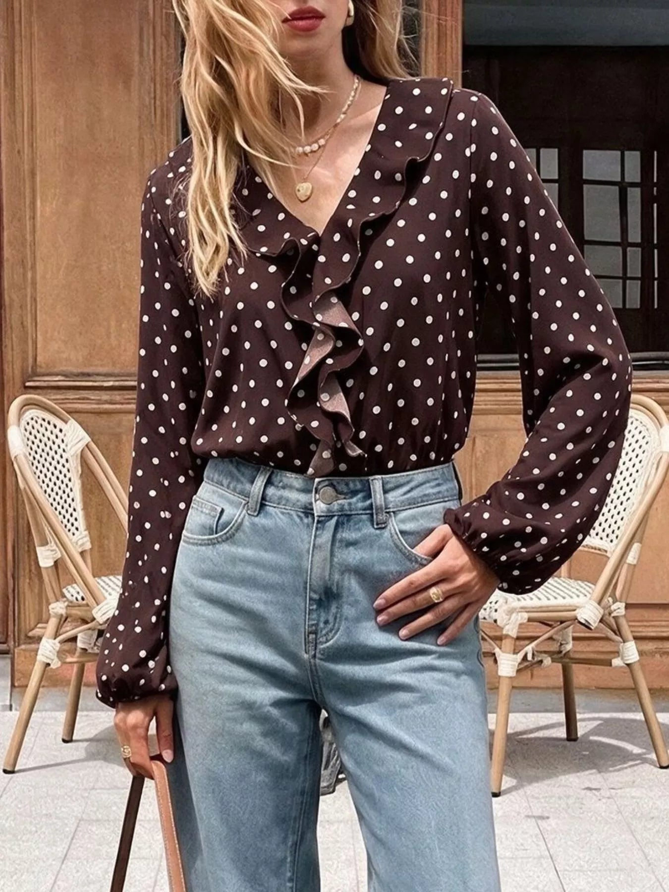 Outdoor front view of a coffee brown polka dot bodysuit with a ruffled V neckline, hand in pocket