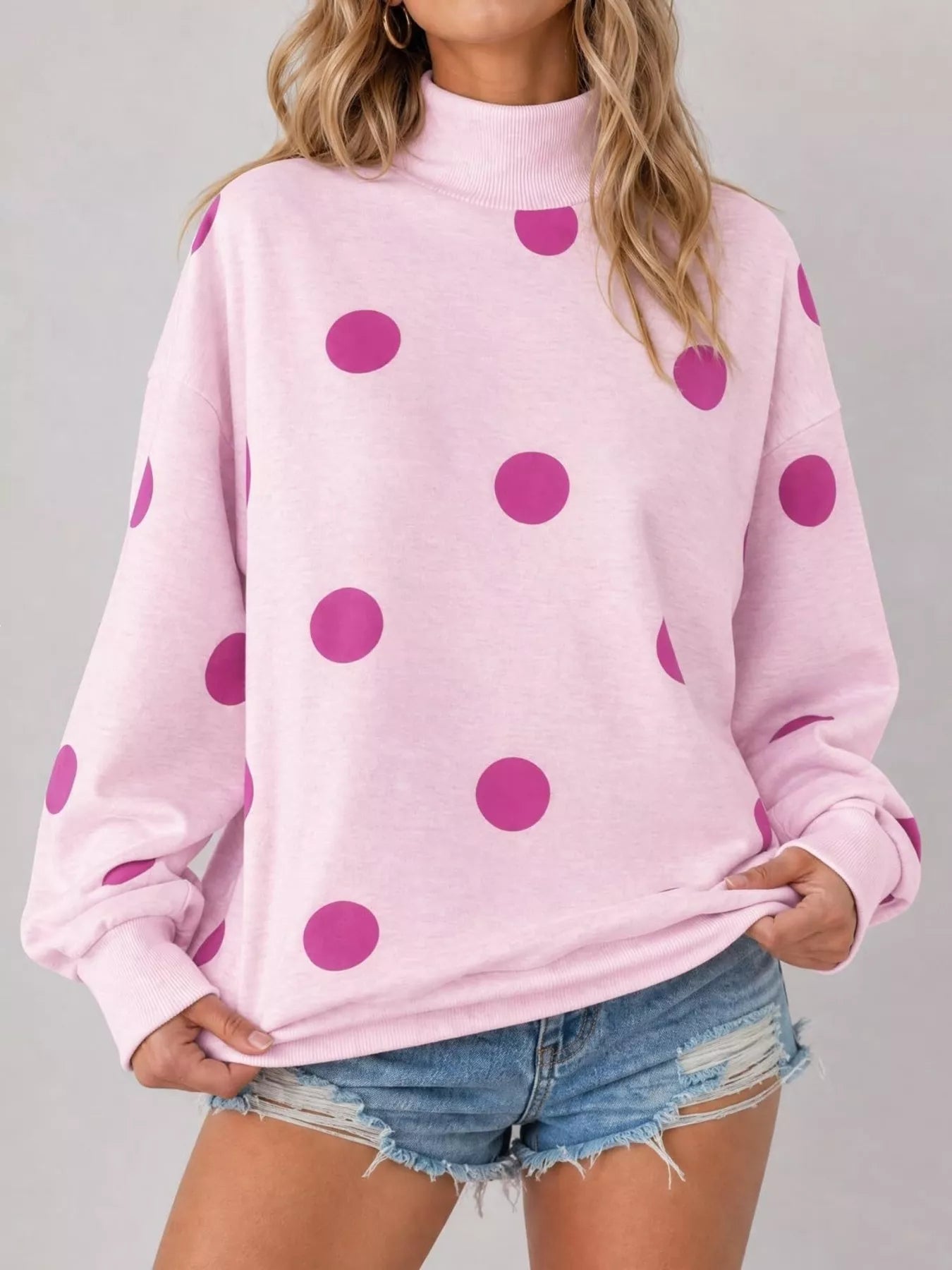 Front studio view of pink mock neck oversized sweatshirt with magenta polka dots holding hem