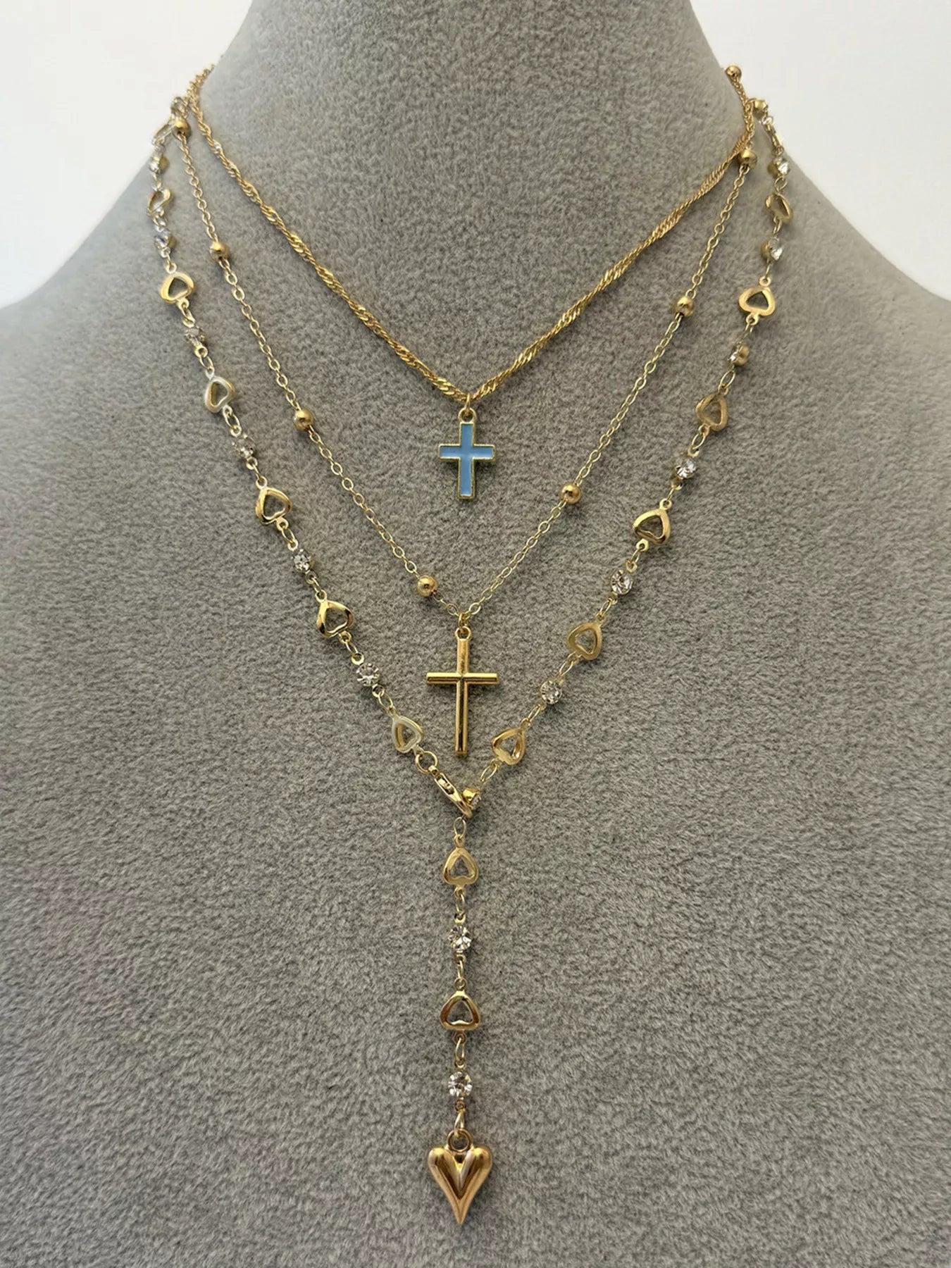 Layered gold necklaces displayed on grey mannequin with blue cross and heart drop