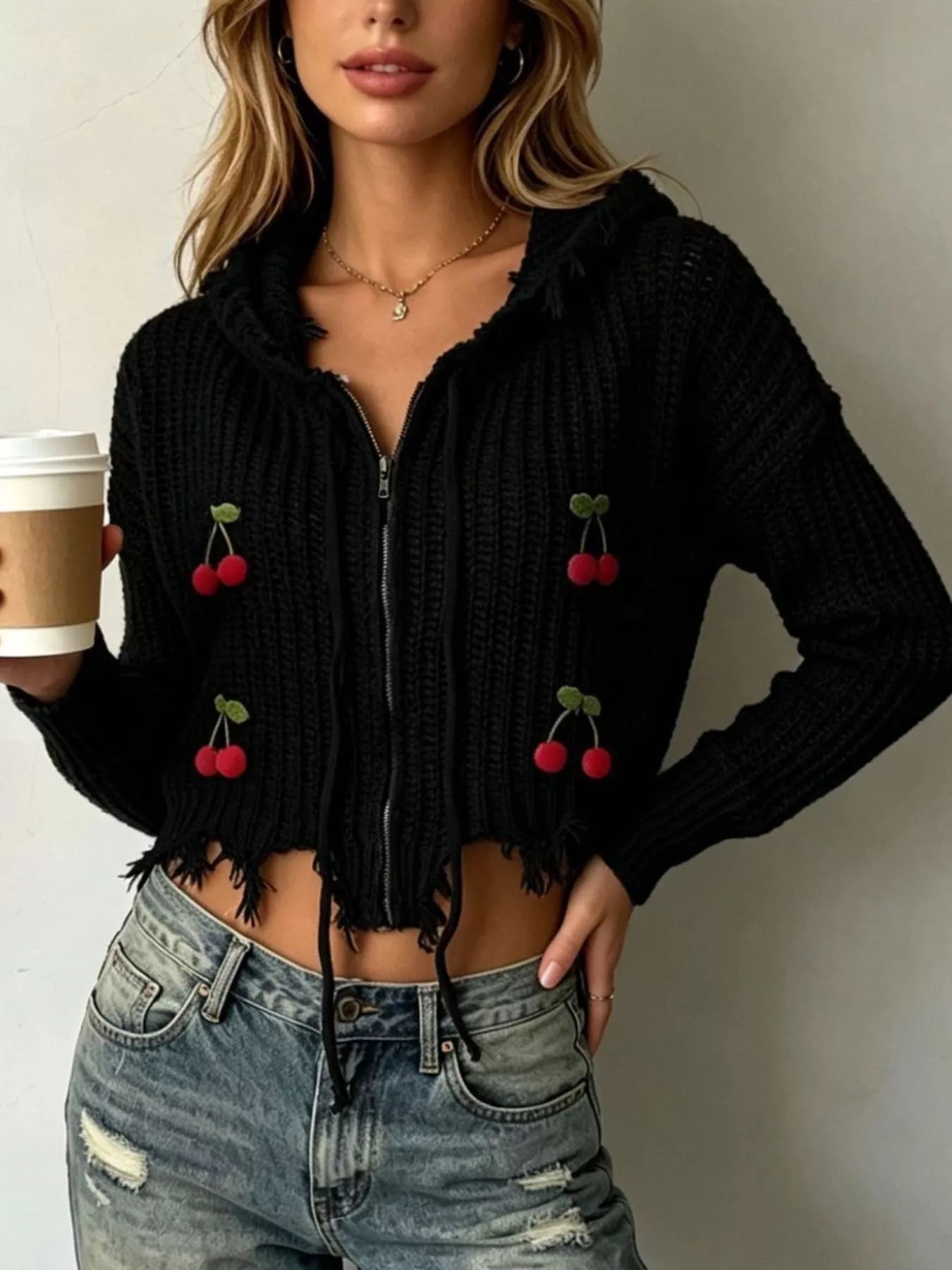 Black knit zip up hooded sweater with red cherry appliques worn on model holding coffee