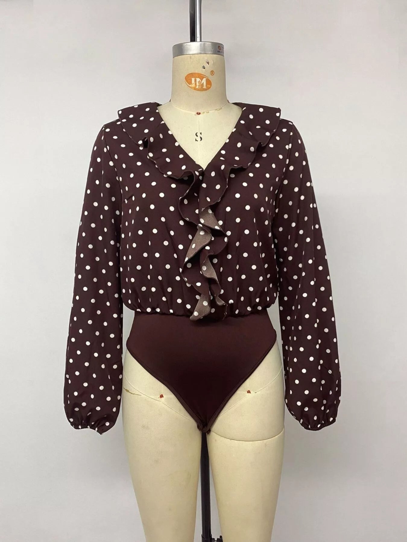 Mannequin front view of a coffee brown polka dot bodysuit with a ruffled V neckline