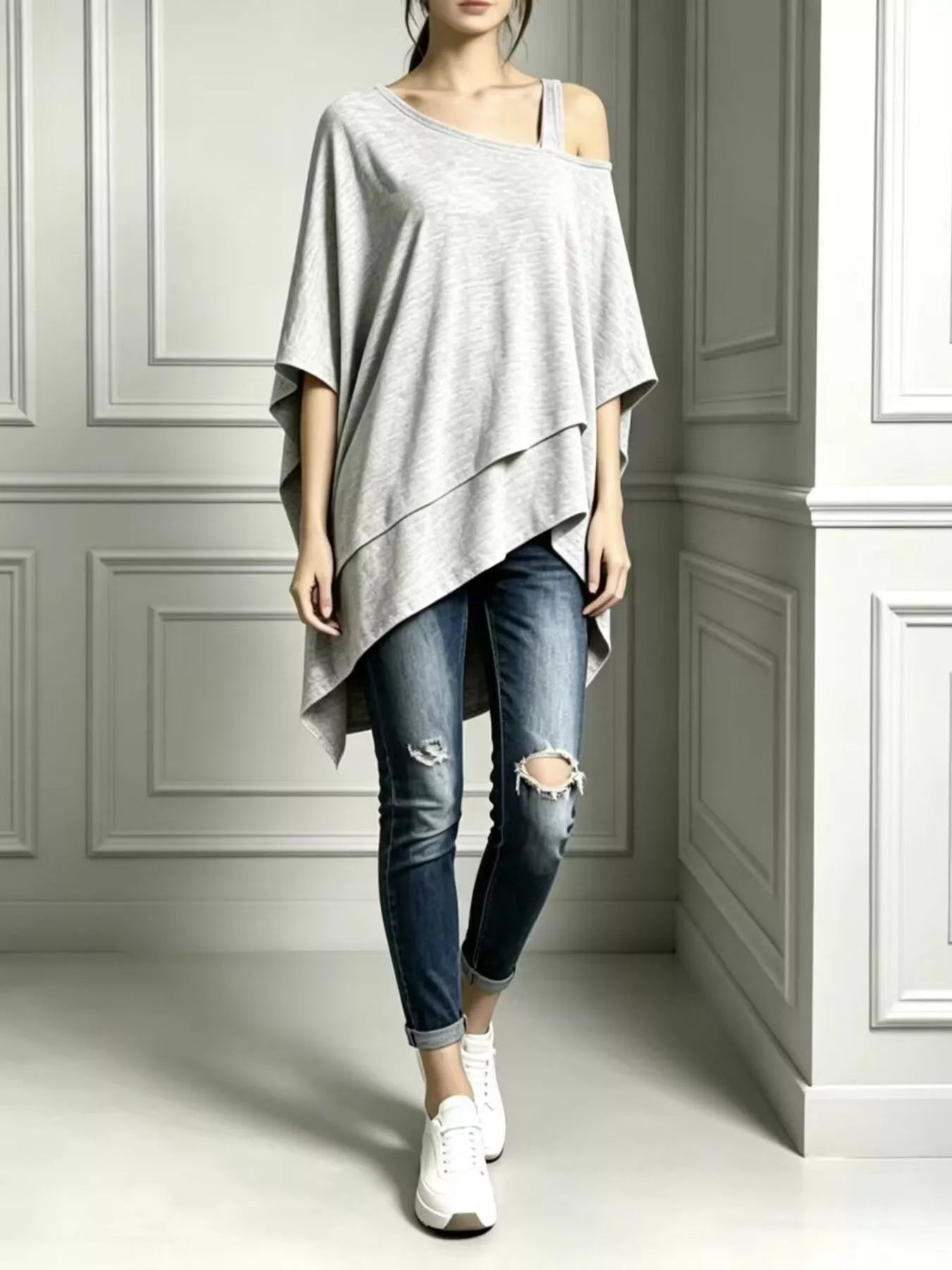 Woman wearing a light gray asymmetrical neck short-sleeve tunic top with distressed jeans and white sneakers.