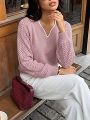 Seated front view of a pink V neck sweater with openwork sleeves, worn with white trousers.