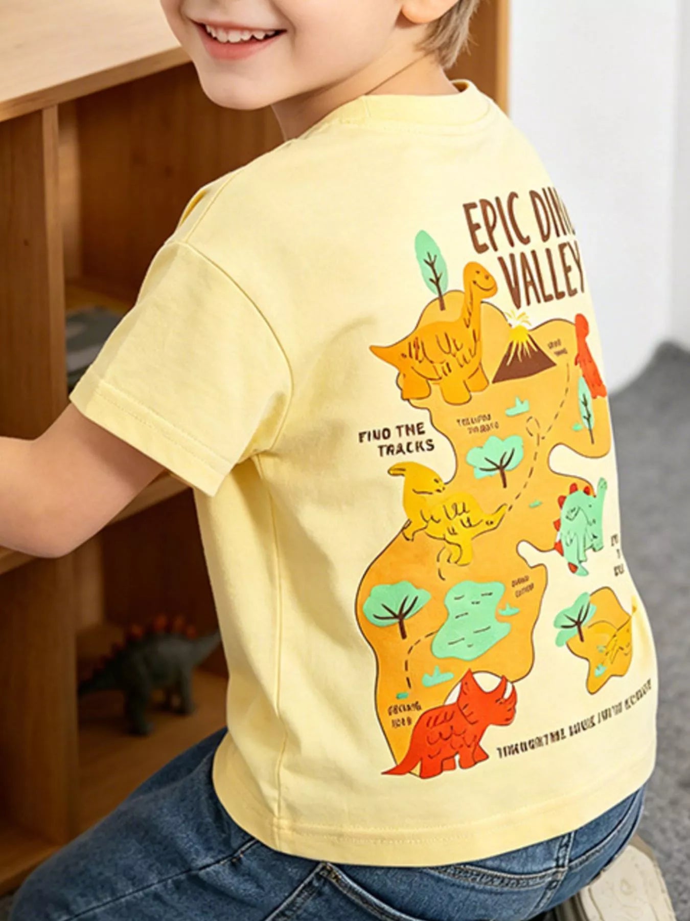 Back view of a pale yellow children's tee with a large dinosaur valley map graphic, worn on a model