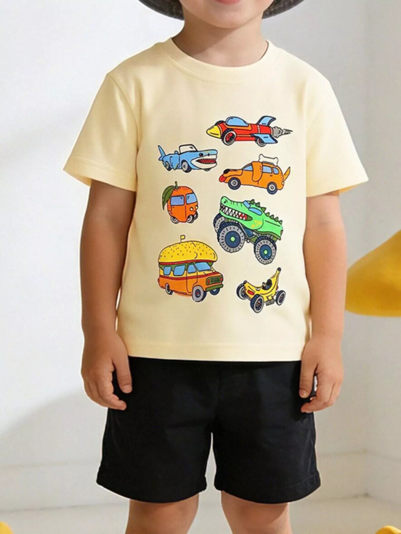 Front view of a cream children's tee with cartoon rocket, shark, dog and monster truck cars