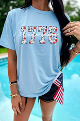 Light blue relaxed T-shirt featuring floral-print “1776” lettering and a white cursive “America” overlay.
