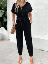 Women's black V-neck jumpsuit with short sleeves, tie detailing, decorative buttons, and pockets.