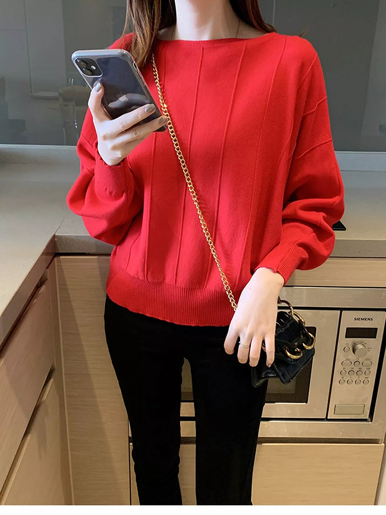 Front view of a red batwing sleeve knit top with vertical seams, holding a phone indoors.