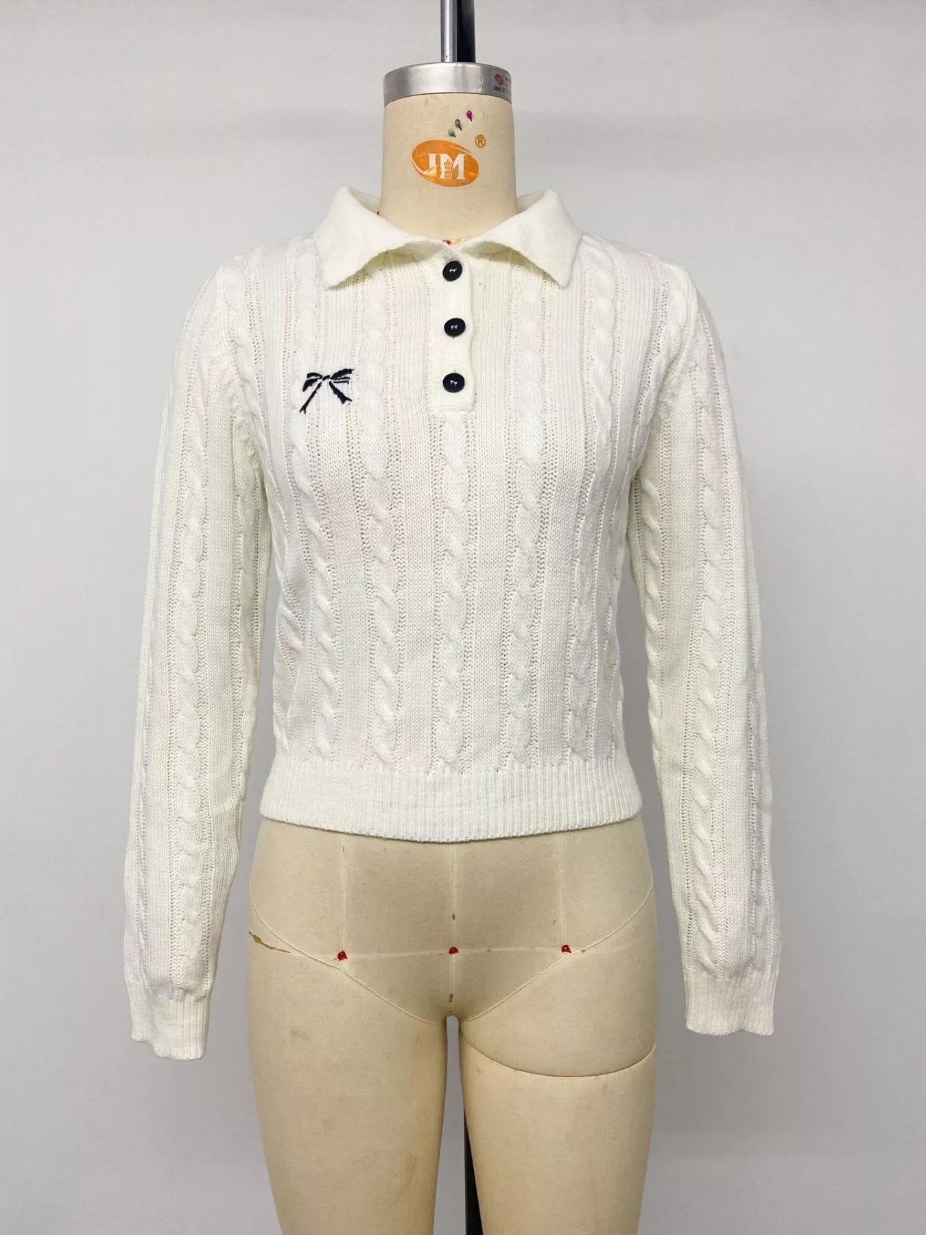 White cable knit polo sweater with black buttons and bow embroidery on mannequin, front