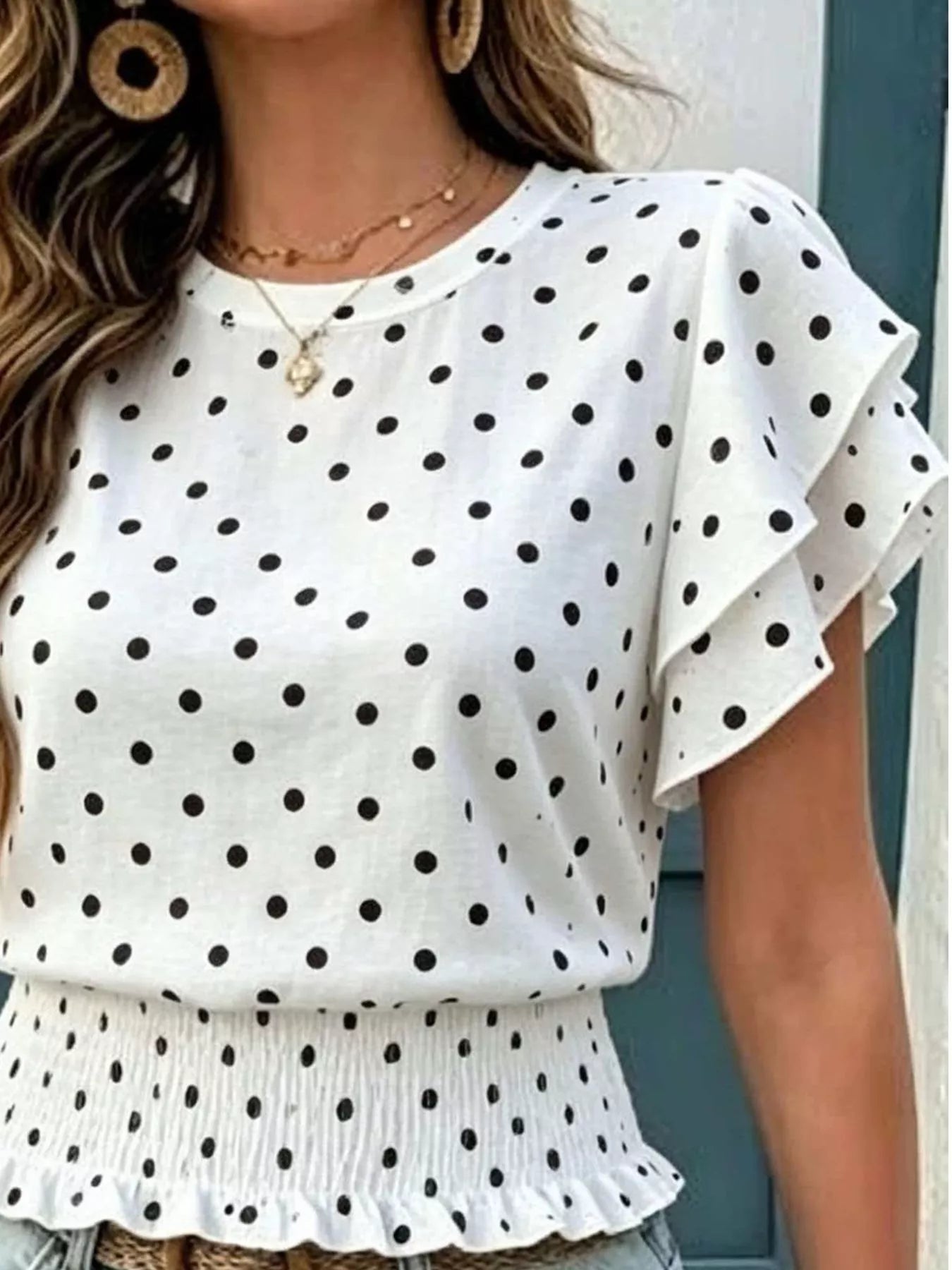 Close-up front view of a white blouse with black polka dots, ruffled sleeves, and a smocked peplum hem