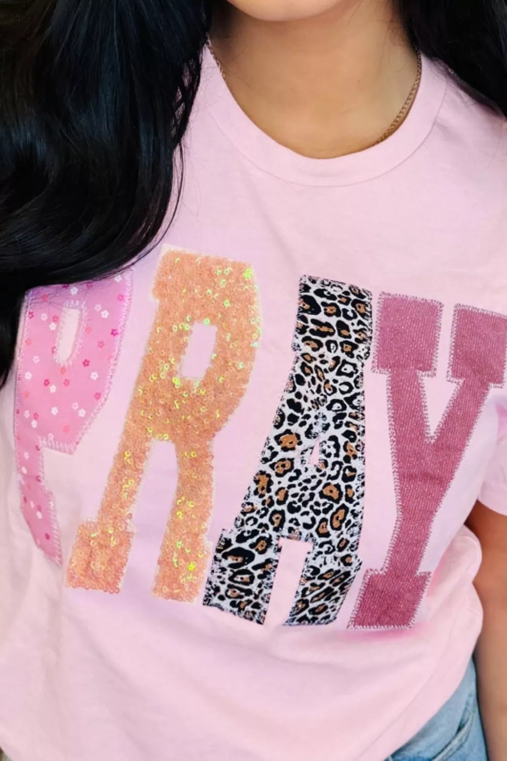 Close-up of pink graphic tee featuring sequined leopard-print patches spelling “PRAY” on the front