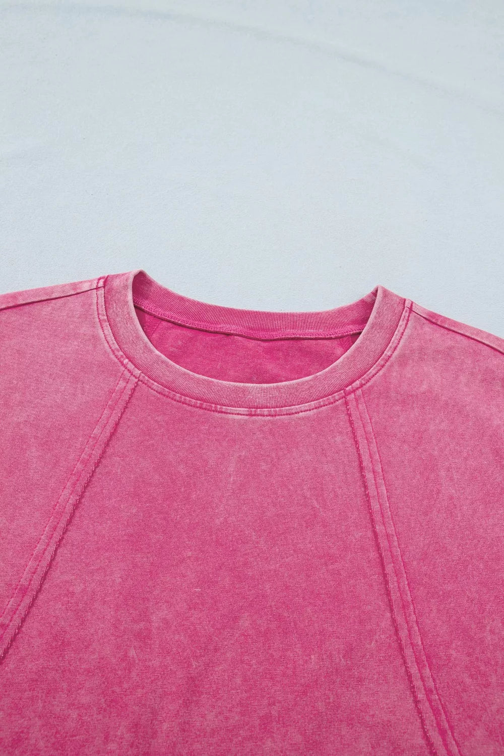 Close-up front detail of a pink mineral-wash top featuring raw seam detailing and a ribbed crew neckline