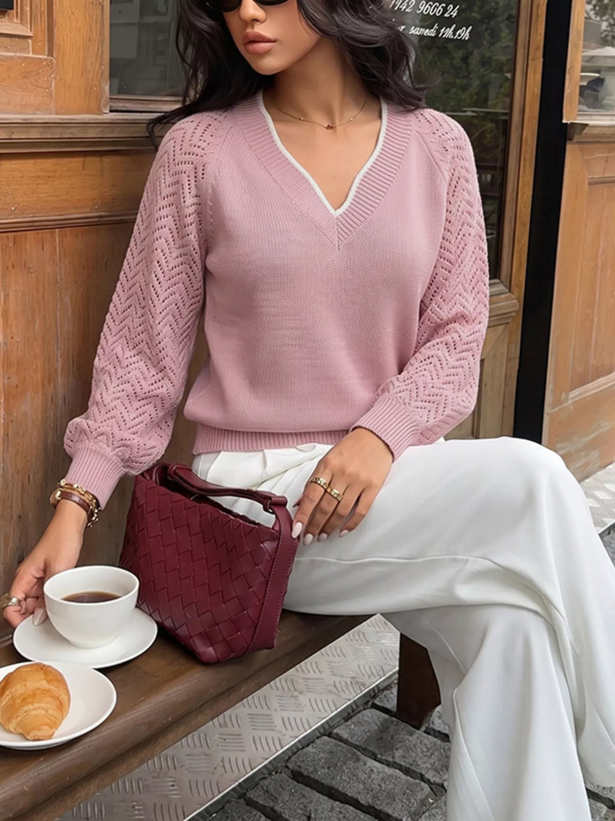 Seated front view of a pink V neck sweater with white trim and openwork chevron long sleeves.