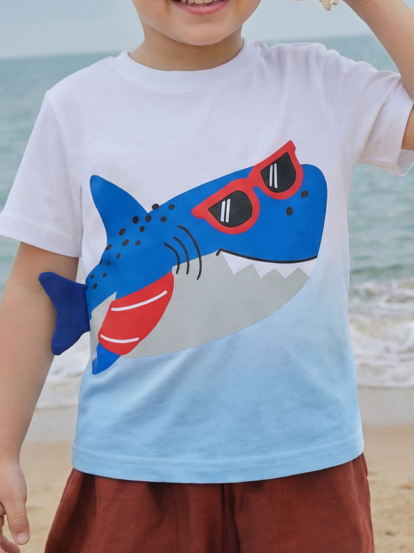 Close-up front view of children's white to light blue T-shirt with a blue shark wearing red sunglasses