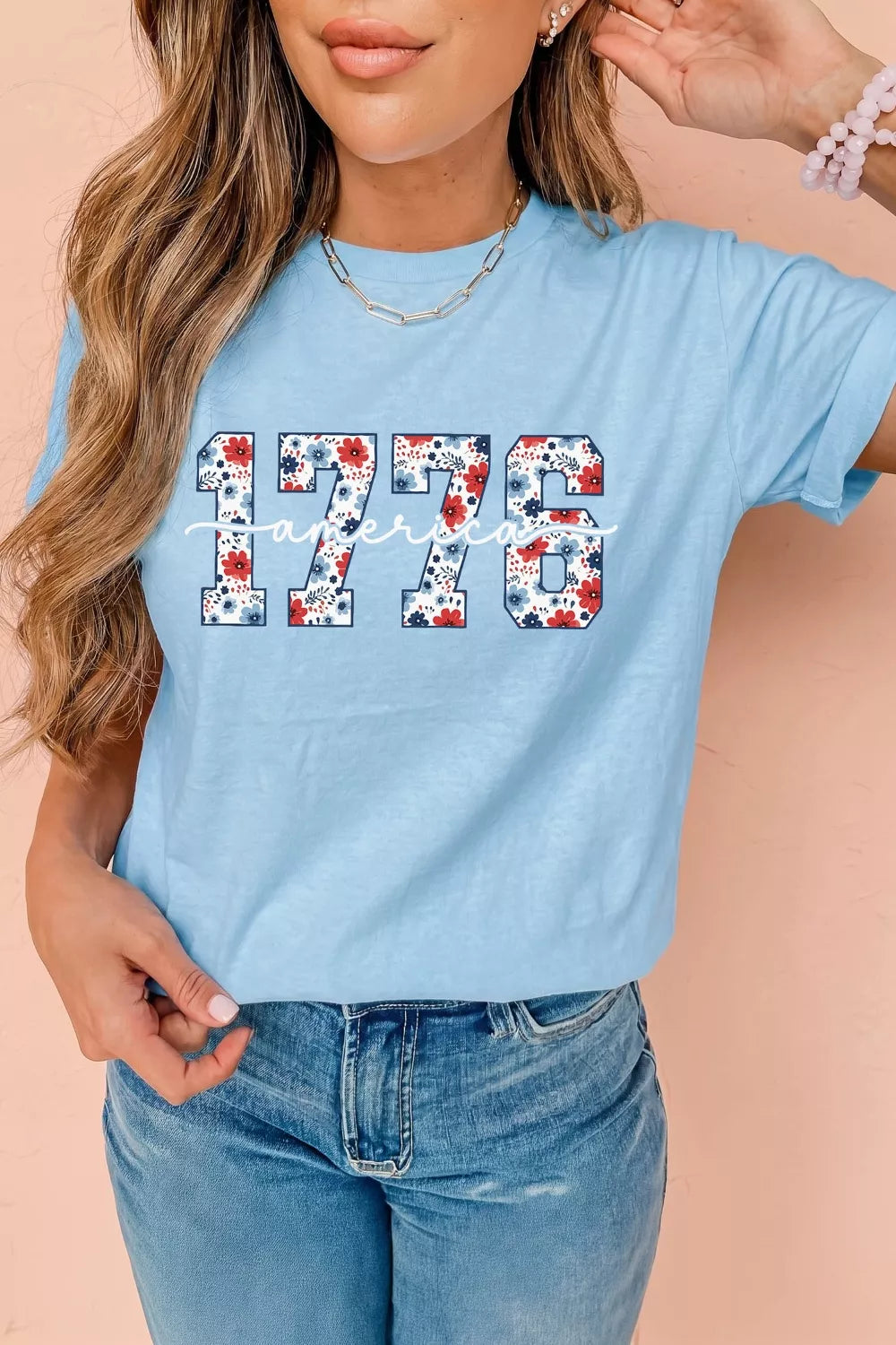 Model wearing a light blue relaxed T-shirt with floral-print “1776” and America script graphic.