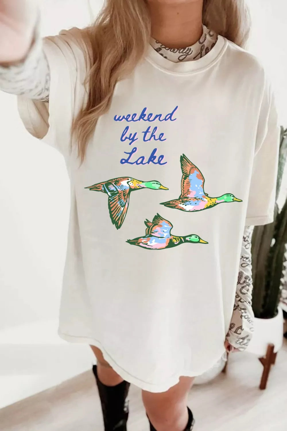 White oversized T-shirt featuring blue “weekend by the Lake” lettering and three colorful flying ducks.