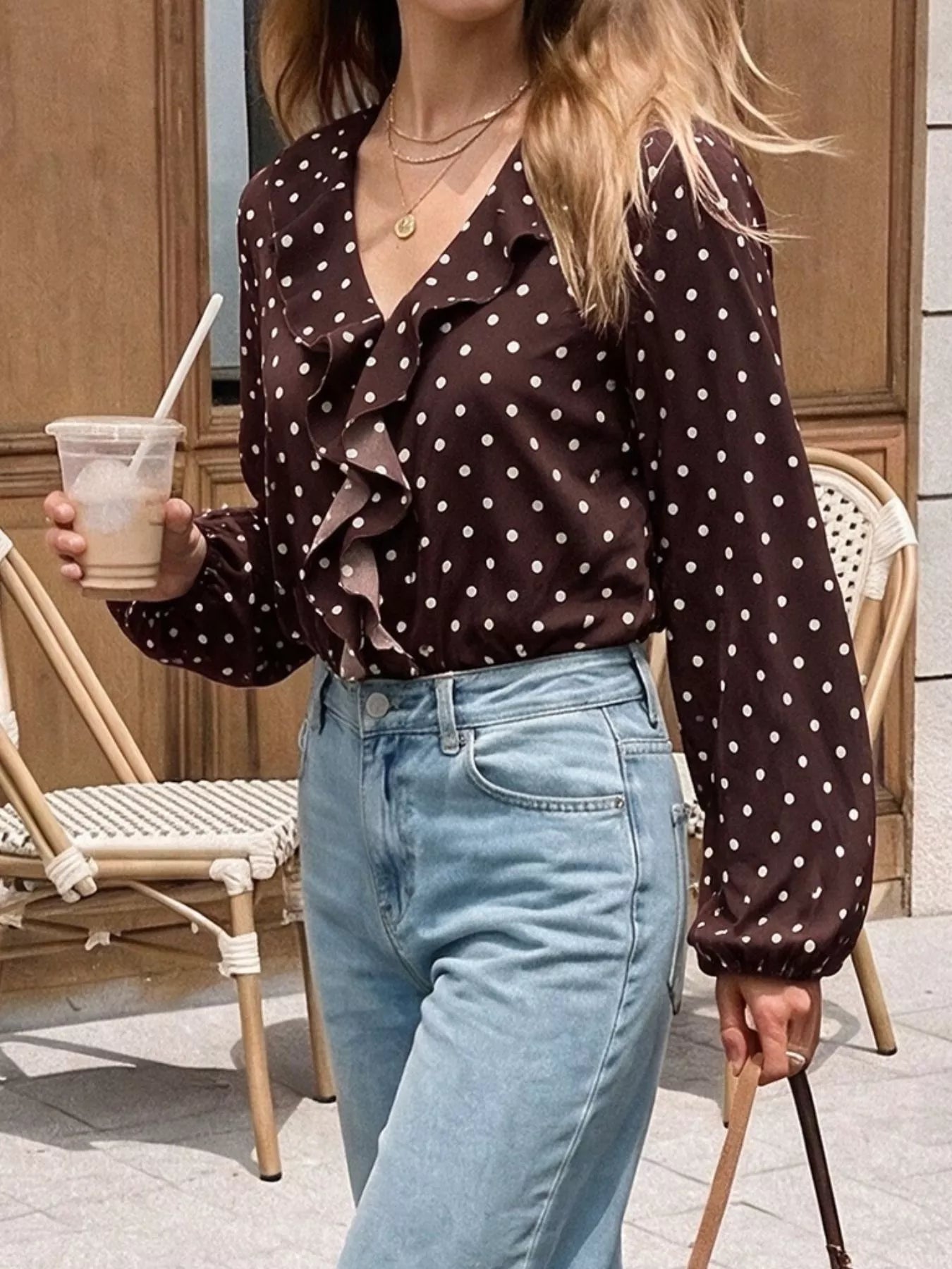 Outdoor standing view of a coffee brown polka dot bodysuit with a ruffled V neckline and long sleeves