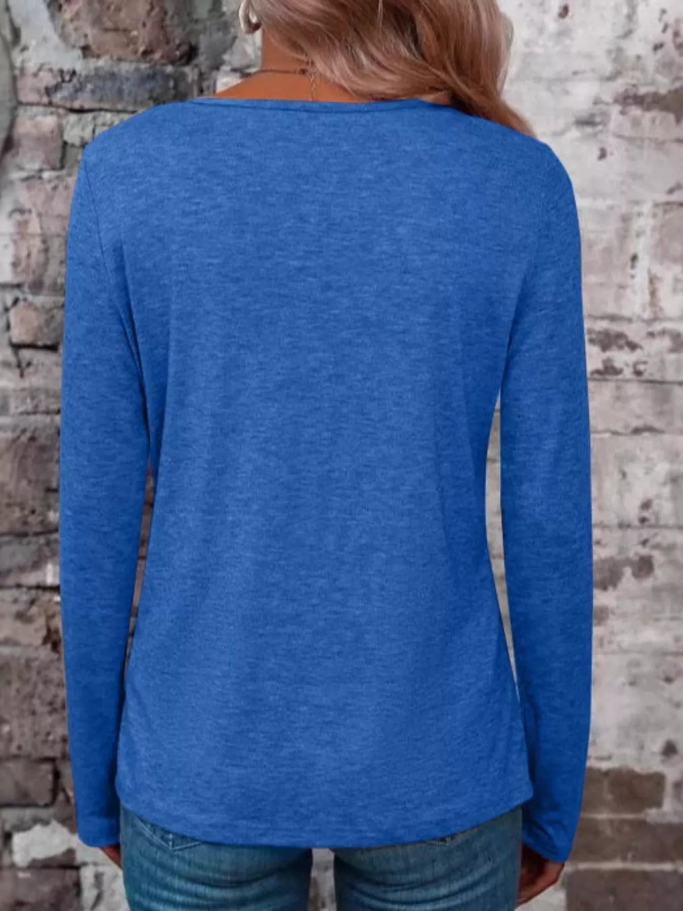 Back view of a blue long-sleeve V-neck top modeled with jeans against a brick wall
