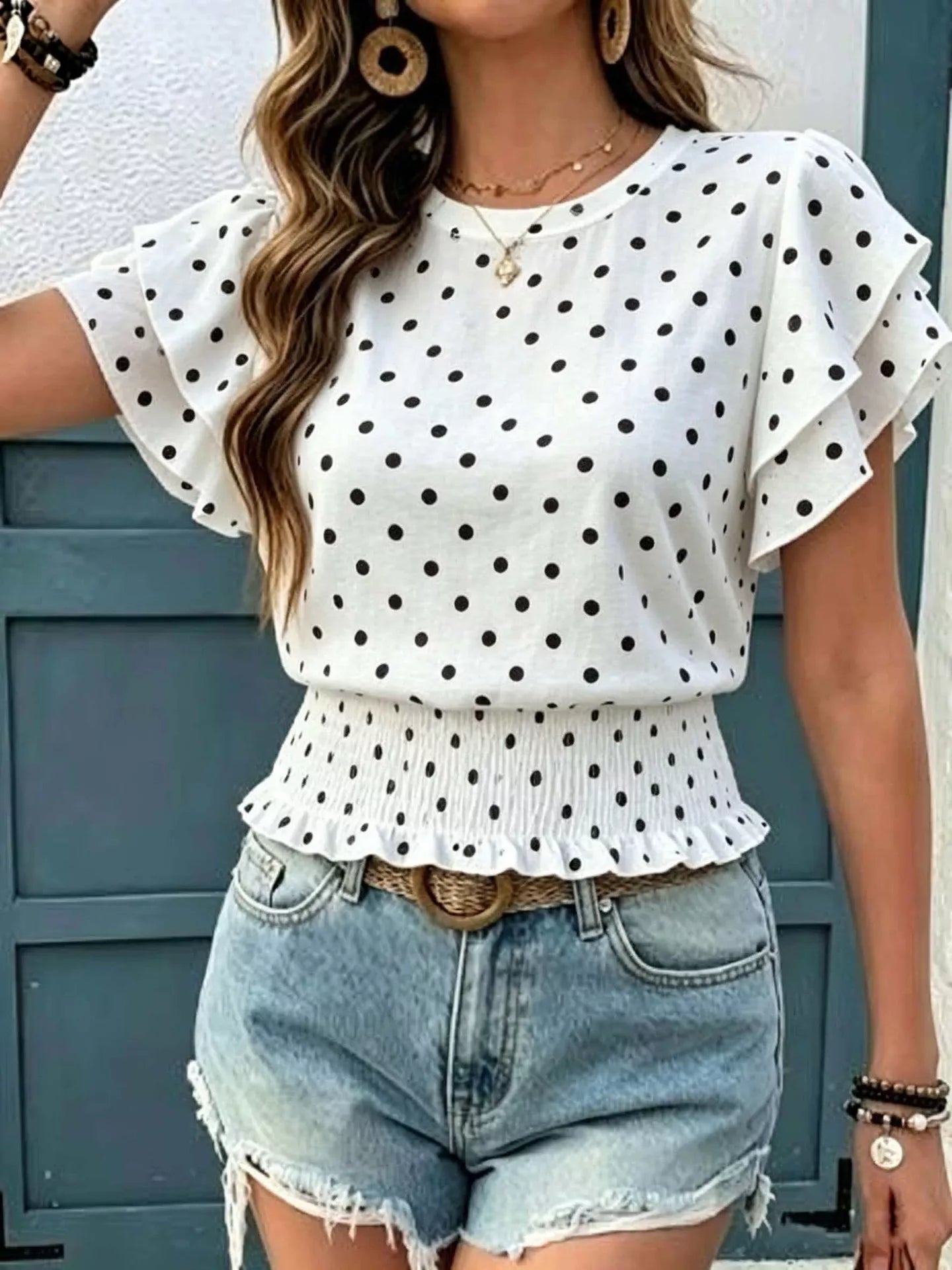White polka dot blouse with ruffled sleeves and smocked peplum waist, shown styled with denim shorts