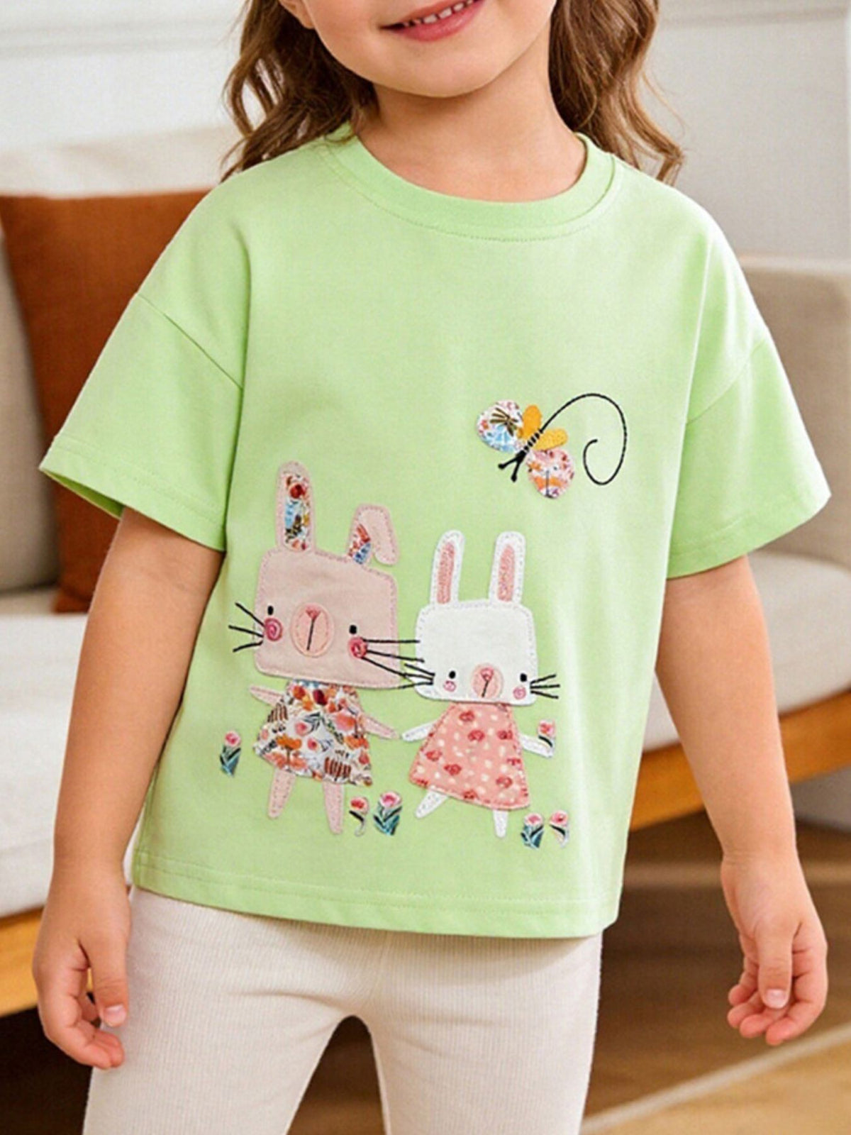 Front view of light green children's tee with two cartoon bunnies in floral dresses and a butterfly