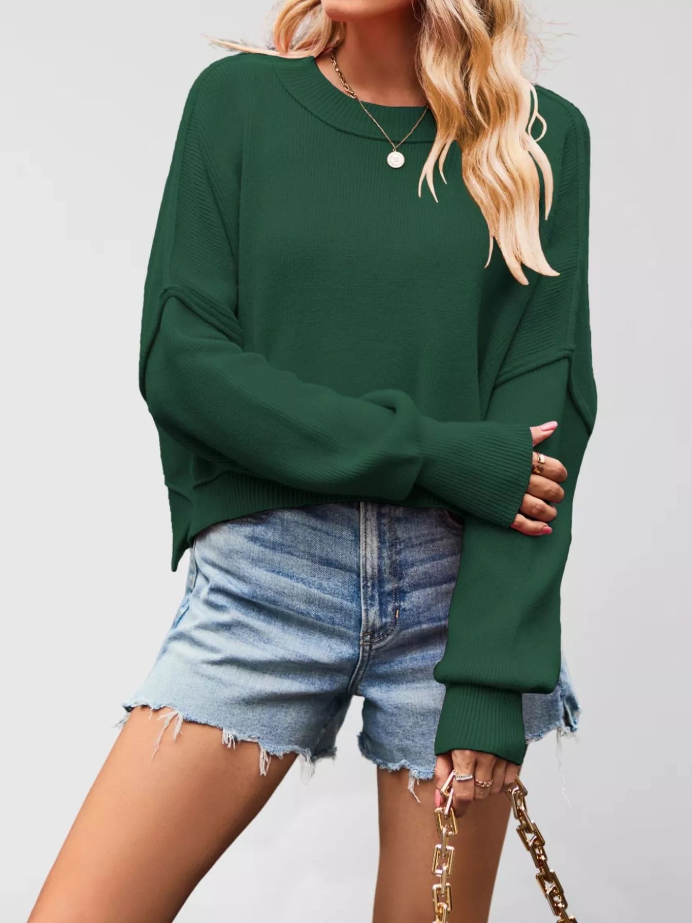 Front view of a dark green crew neck dropped shoulder sweater with ribbed cuffs worn with denim shorts.