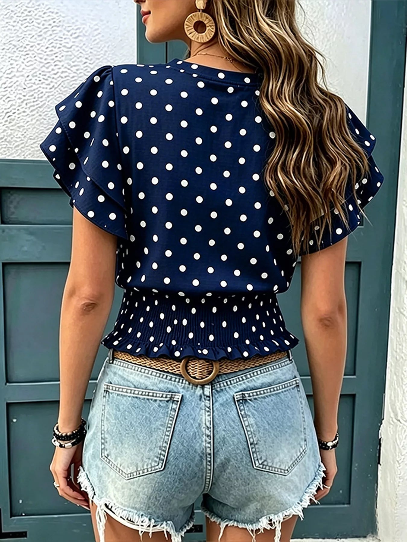 Back view of navy polka dot blouse with ruffle sleeves and peplum hem, worn with denim shorts