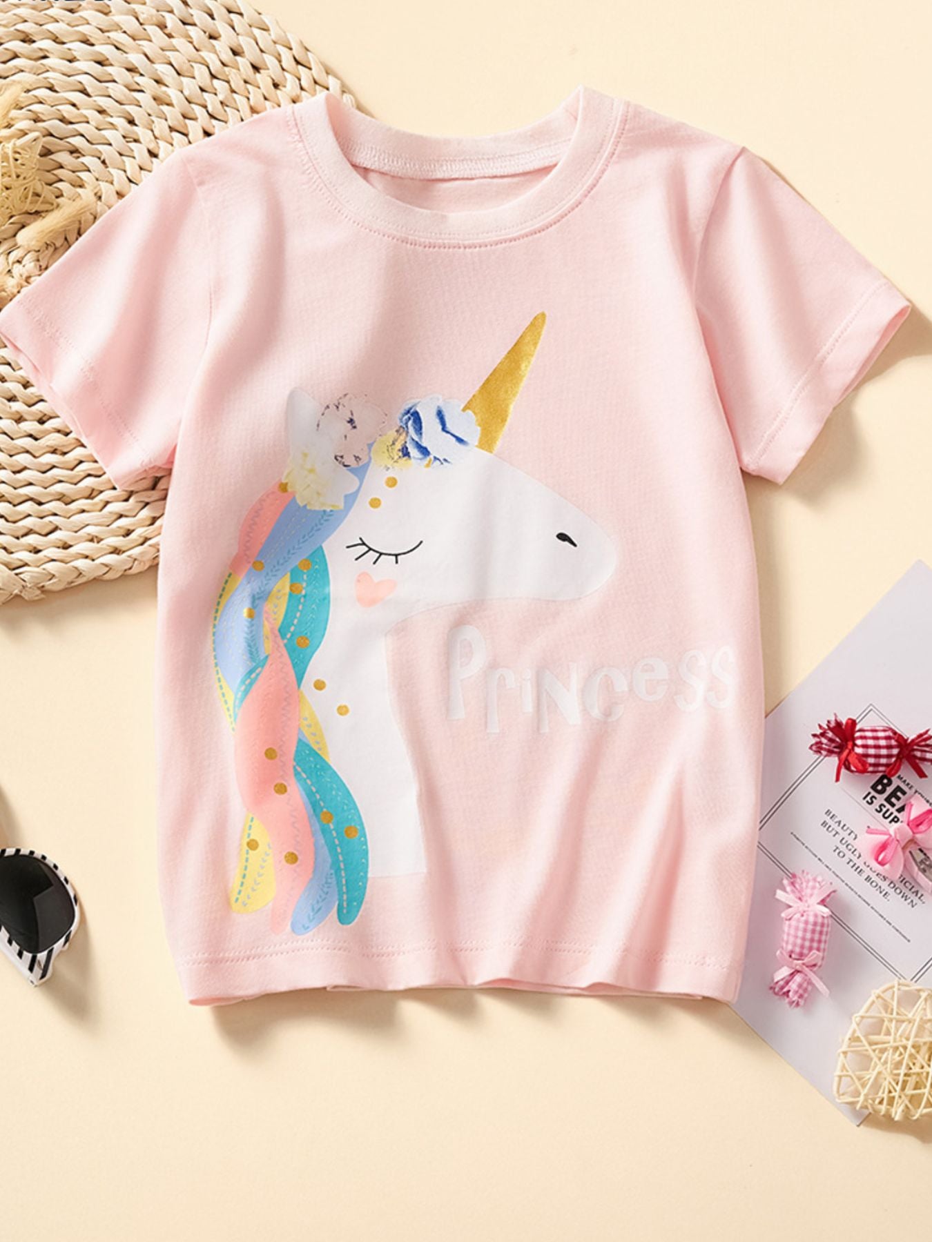 Flat front view of a blush pink children's tee with a white unicorn and rainbow mane