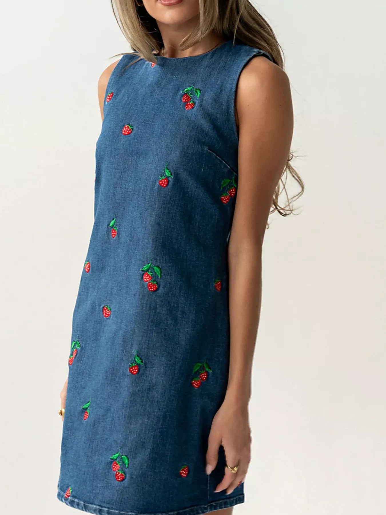 Side view of blue sleeveless denim dress with scattered red strawberry embroidery