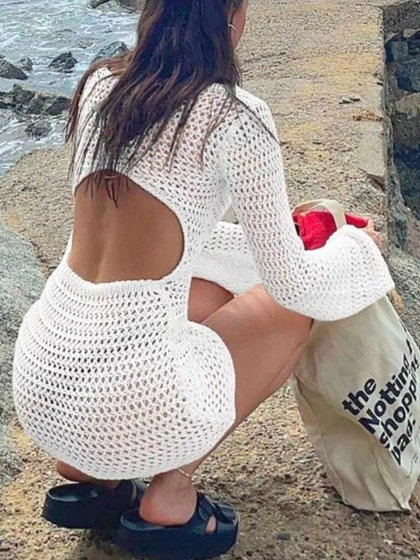 Back view of white crochet dress with large back cutout, model crouching by shore