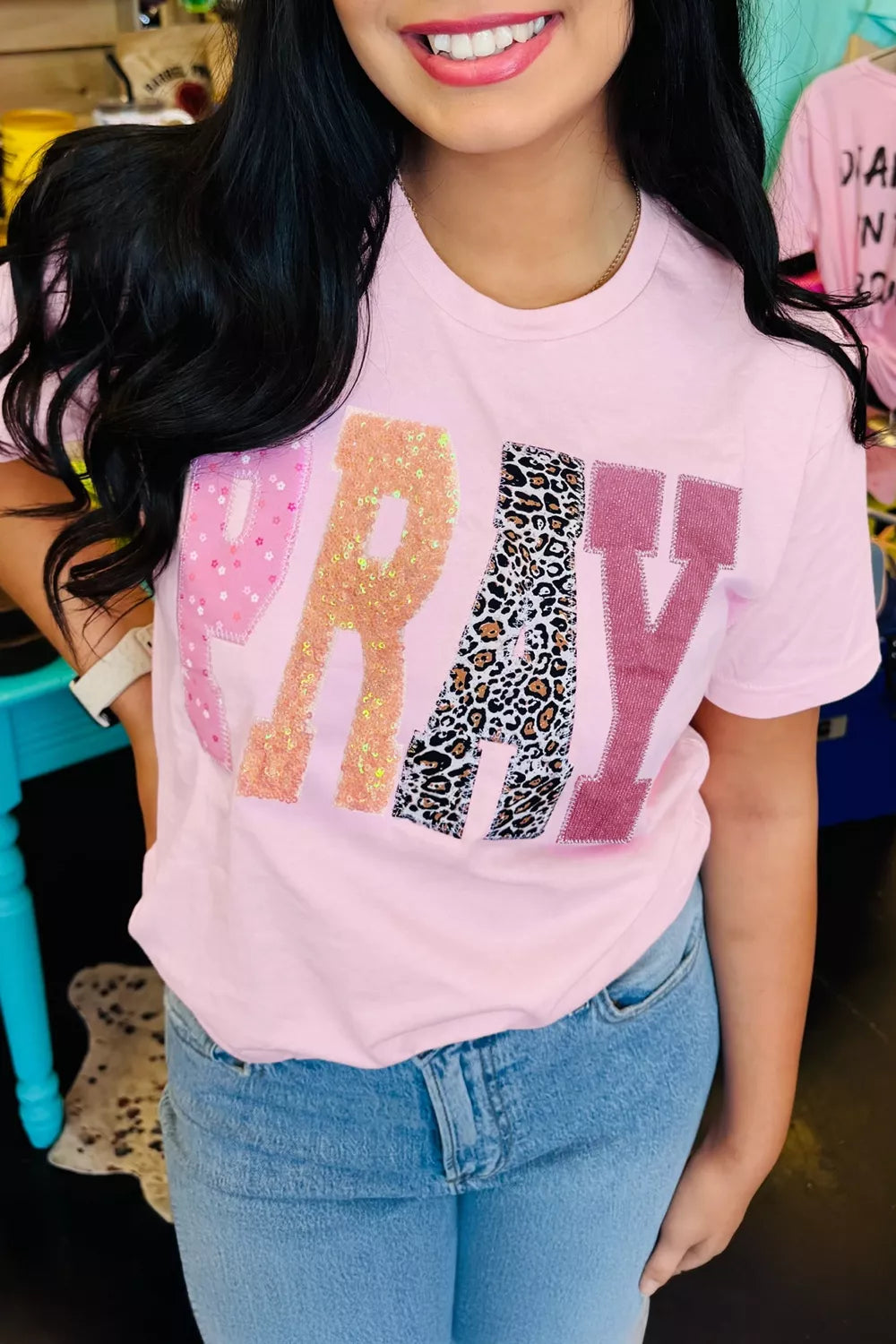 Pink PRAY graphic tee with sequin, leopard-print, and patterned letter patches, shown worn with jeans