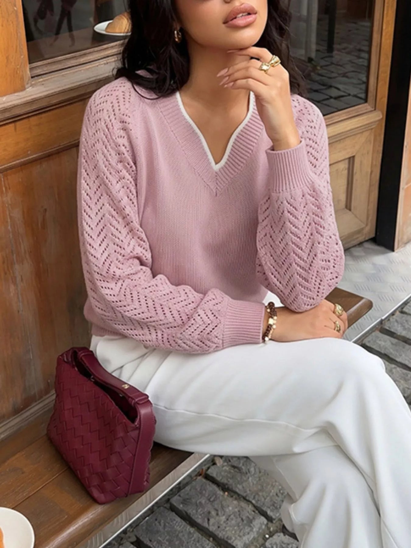 Seated front view of a pink V neck sweater with openwork sleeves, worn with white trousers.