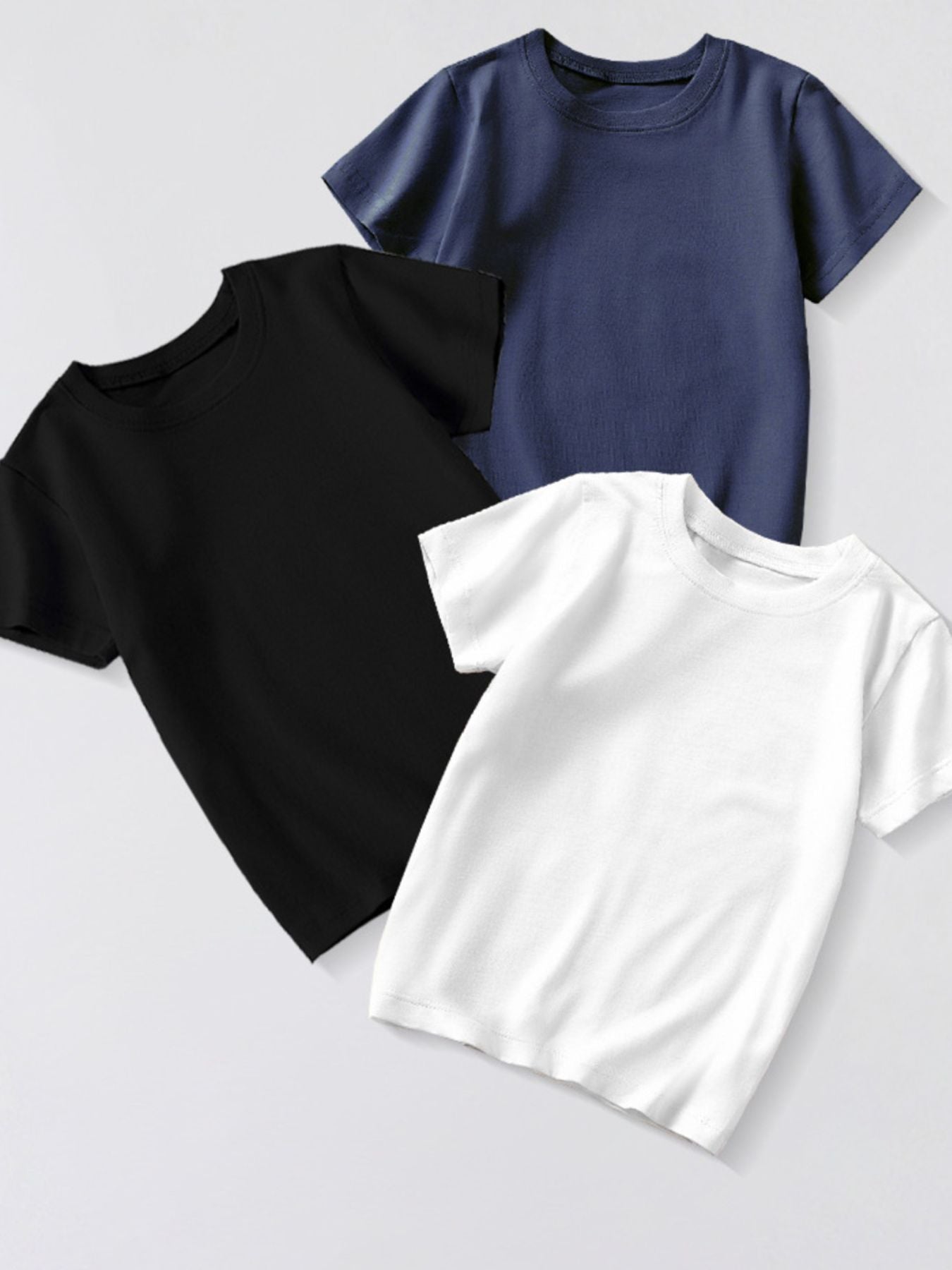 Three plain children's crew neck T-shirts in dark navy, black and white, arranged overlapping in a flat lay