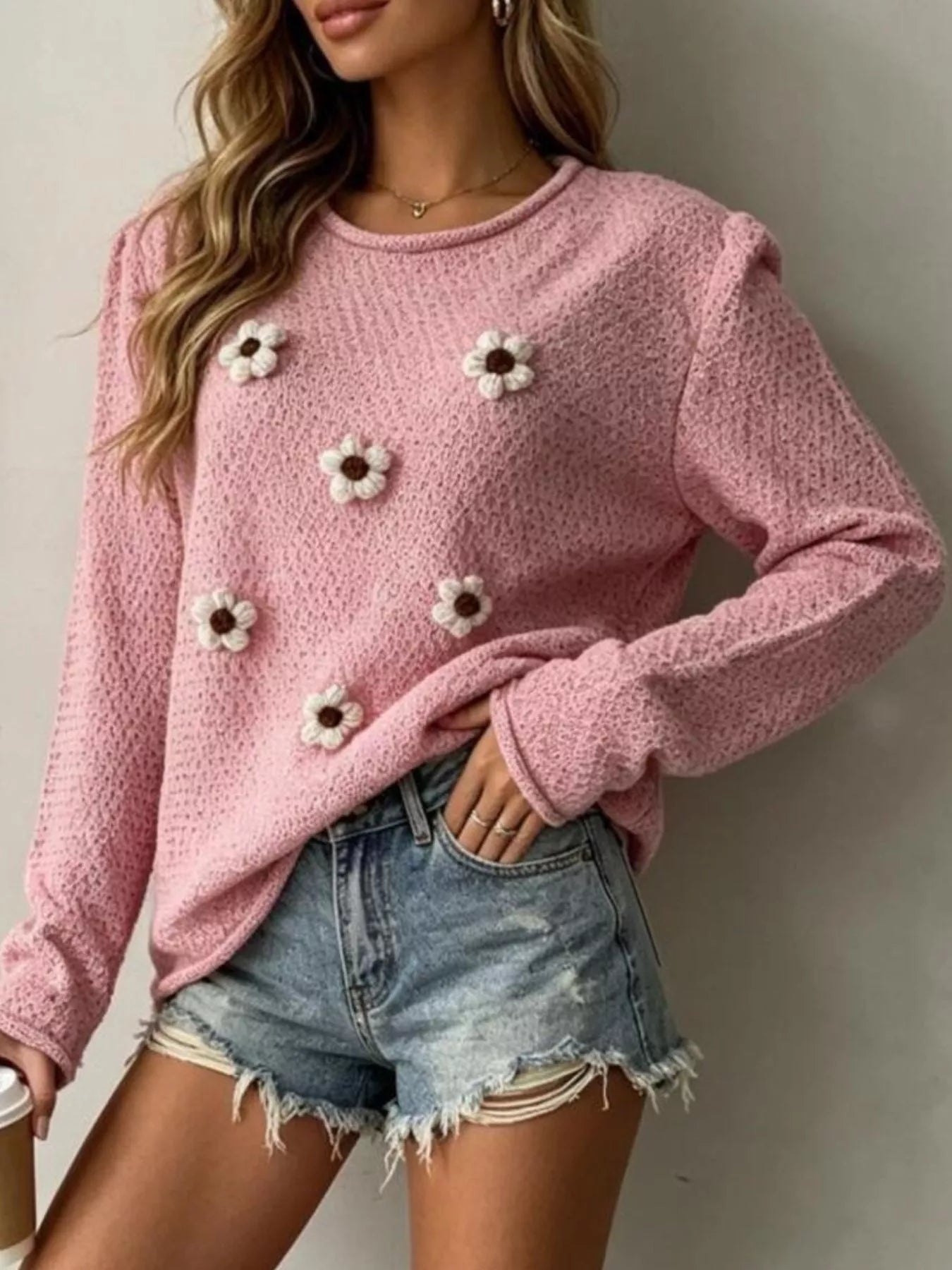 Seated view of a pink textured knit sweater with white floral appliques and long sleeves