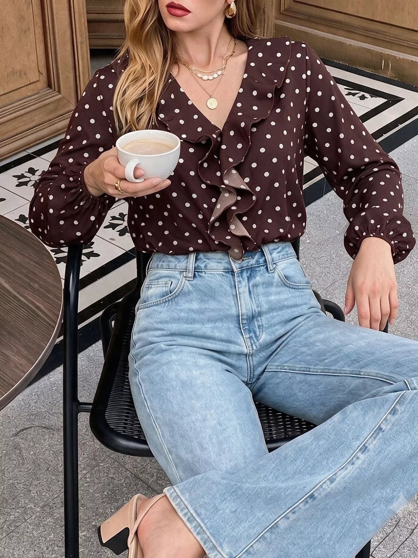 Seated outdoor view of a coffee brown polka dot bodysuit with a ruffled V neckline and long sleeves