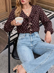Seated outdoor view of a coffee brown polka dot bodysuit with a ruffled V neckline and long sleeves