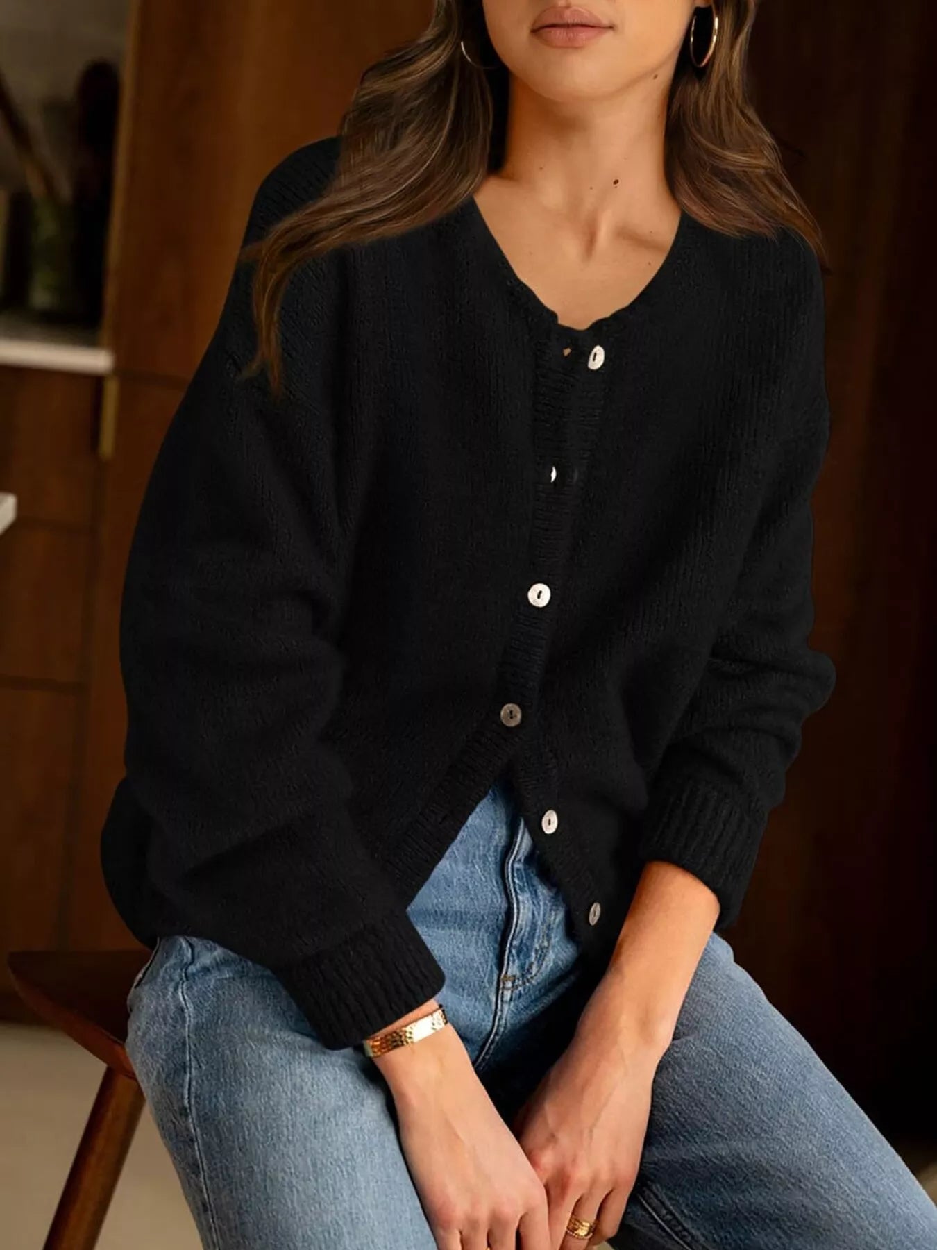 Seated front view of black button down cardigan with white buttons and blue jeans