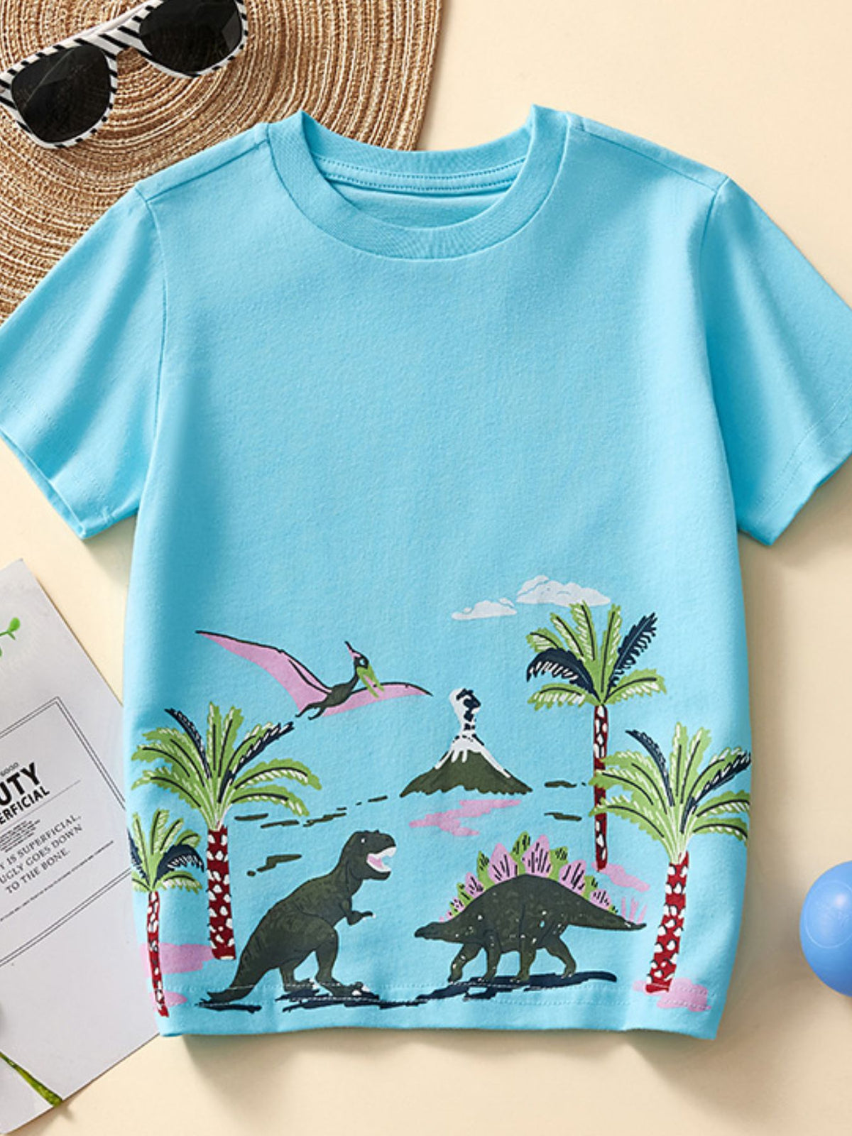 Flat lay front view of pastel blue children's tee with dinosaurs, palm trees and a volcano print