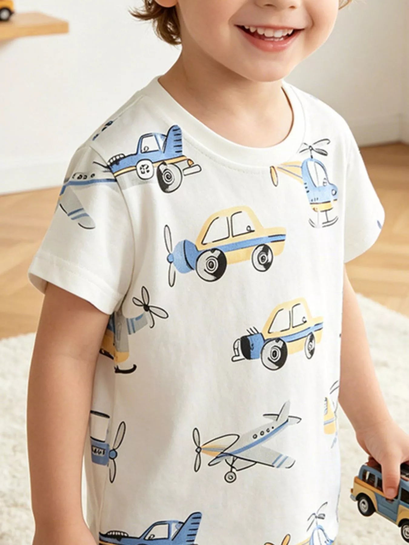 White children's cotton t-shirt with cars, helicopters, and airplanes, shown worn indoors