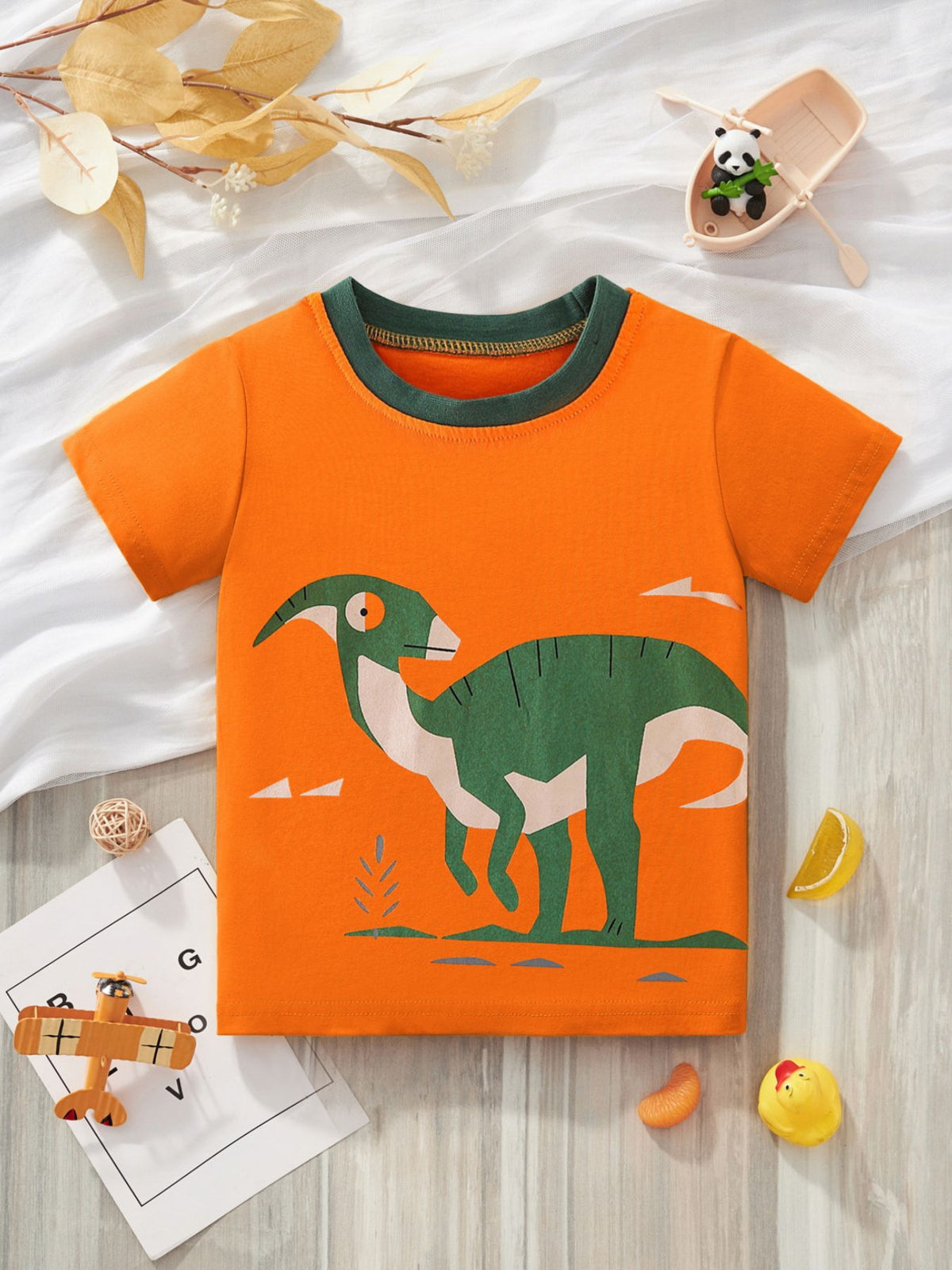 Orange children's crew neck T-shirt with a large green dinosaur graphic, laid flat among small props