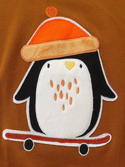 Detail view of the penguin applique wearing an orange hat and riding a red skateboard on a caramel sweatshirt