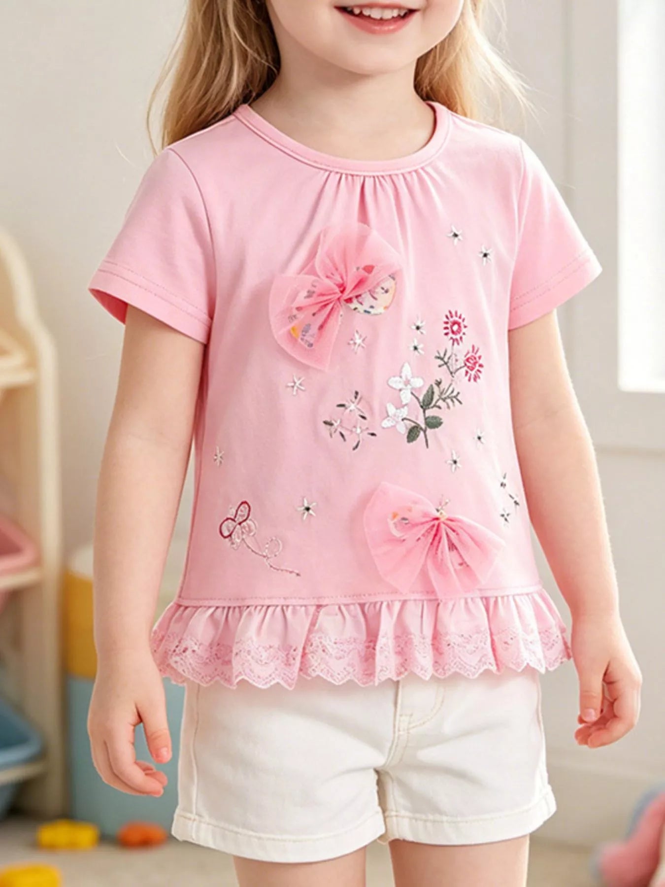 Front view of a children's blush pink t-shirt with tulle bows, floral embroidery and a ruffled lace hem