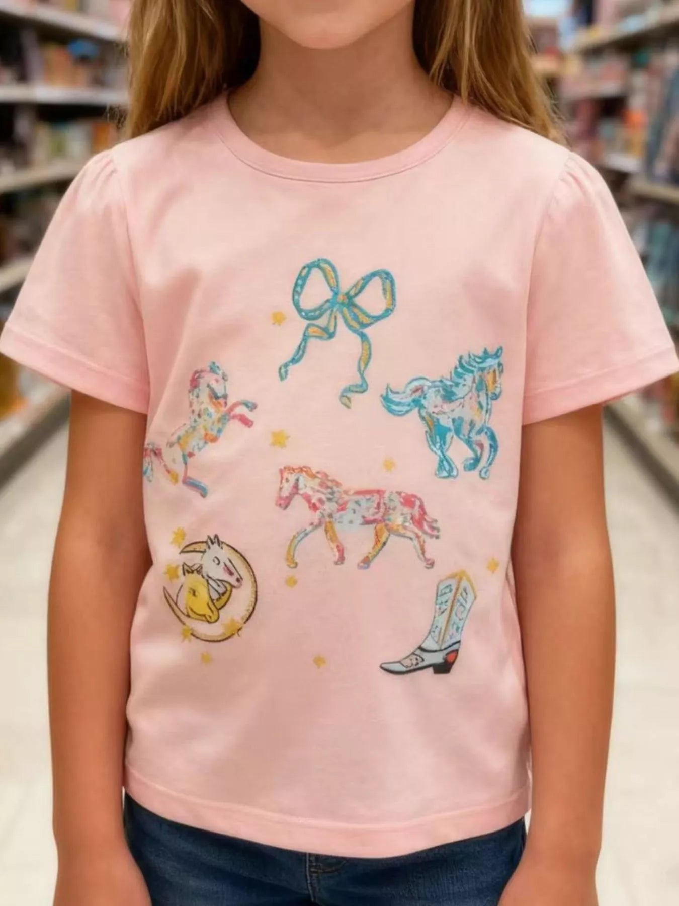 Front view of a pink children's tee with colourful horses a bow a cowboy boot and stars